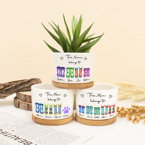 Könnte beinhalten: Drei weiße Keramik-Pflanztöpfe mit Bambus-Untersetzern. Jeder Topf ist mit bunten Gummistiefeln und den Texten "This Nanny belongs to" oder "This Mum belongs to" mit Namen verziert.