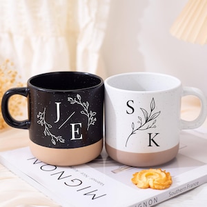 Puede incluir: Dos tazas de cerámica, una negra y otra blanca, con acabados moteados y bases color canela. La taza negra presenta las iniciales "J" y "E" con un diseño floral, mientras que la taza blanca muestra "S" y "K" con un motivo botánico similar. Una pequeña galleta descansa cerca.