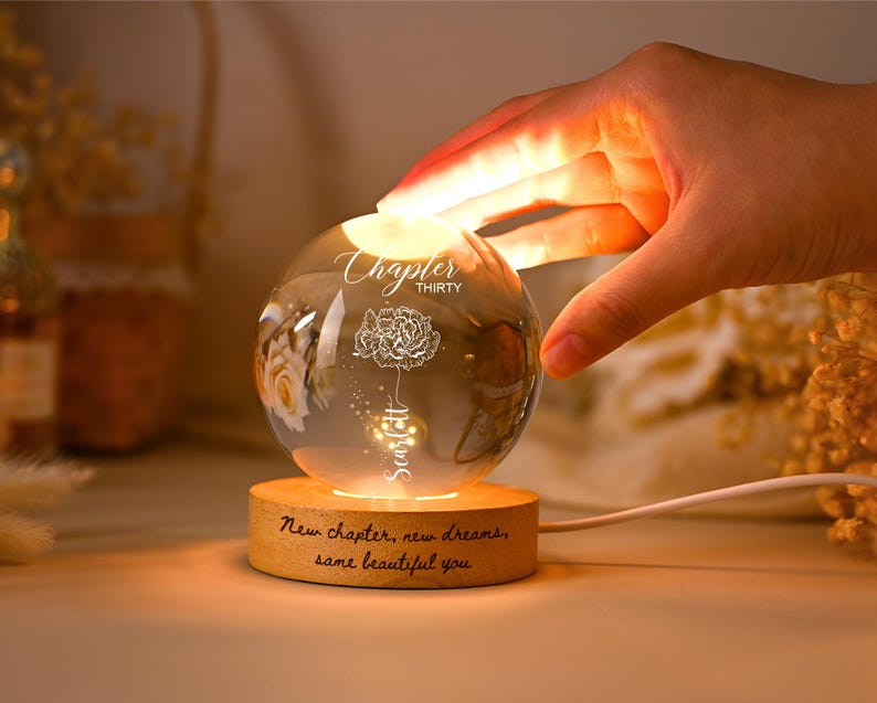 May include: A lit glass sphere with a floral design and the text "Chapter Thirty" and "Scarlett". The sphere is on a wooden base inscribed with "New chapter, new dreams, same beautiful you".