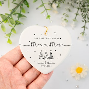 May include: White heart-shaped ceramic ornament with black text that reads "Our first Christmas as Mr & Mrs. Kent & Alice 09.21.2024" and a drawing of three Christmas trees.