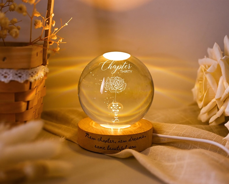 May include: A glowing glass sphere with a floral design and the words "Chapter Thirty" and "Sanjit" etched on it. The sphere sits on a wooden base with the inscription "New chapter, new dreams, same beautiful you."