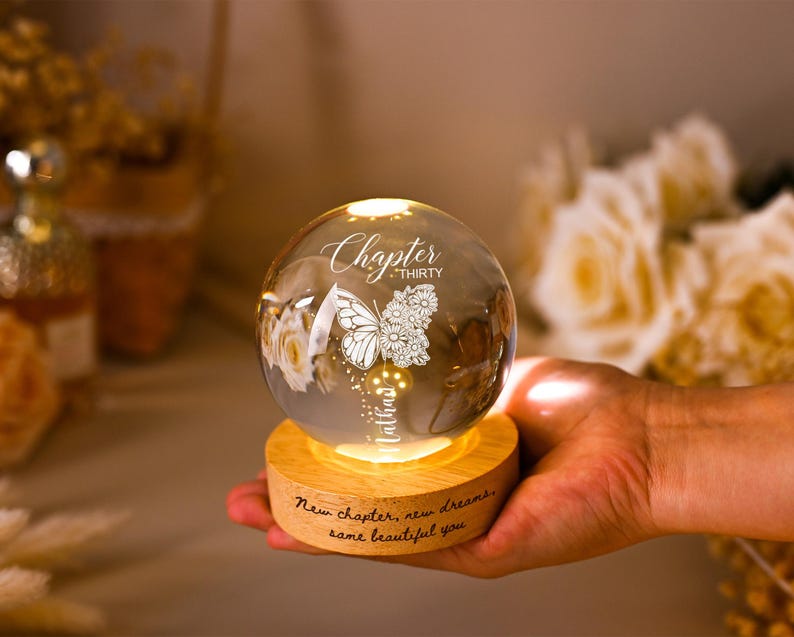 May include: A glowing glass sphere with a butterfly and floral design, resting on a wooden base. The sphere has the words "Chapter Thirty" and "Nathan" etched on it. The base reads "New chapter, new dreams, same beautiful you."