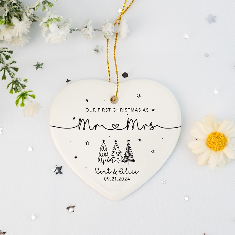 May include: White heart-shaped ceramic ornament with black text that reads "Our first Christmas as Mr & Mrs Kent & Alice 09.21.2024". The ornament features a stylized drawing of three Christmas trees.