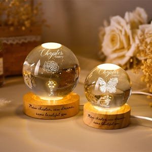 May include: Two illuminated glass spheres with floral and butterfly designs sit on wooden bases. The spheres have the text "Chapter Thirty" and "New chapter, new dreams, same beautiful you" engraved on them. A decorative item.