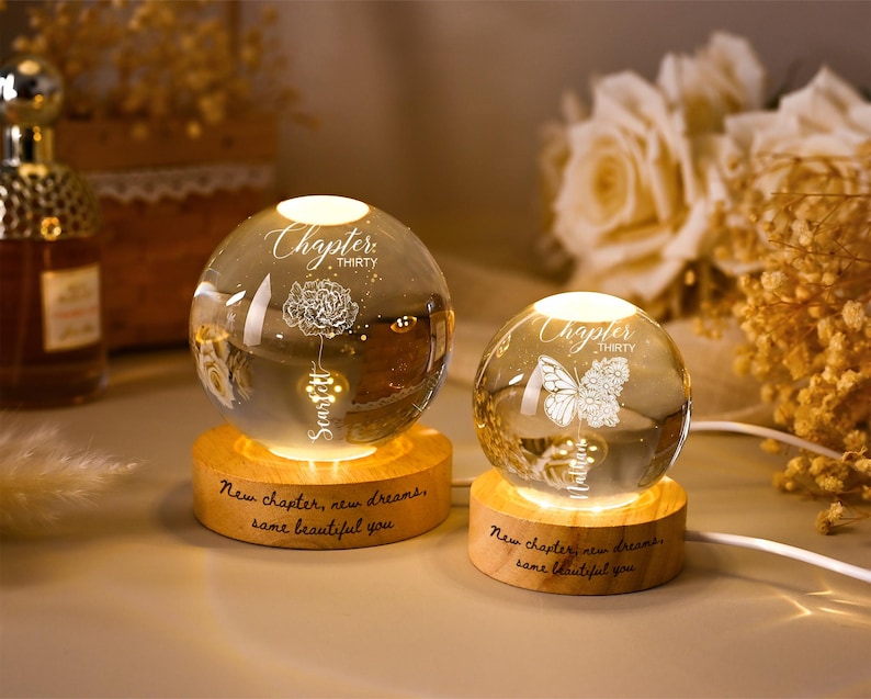 May include: Two illuminated glass spheres with floral and butterfly designs sit on wooden bases. The spheres have the text "Chapter Thirty" and "New chapter, new dreams, same beautiful you" engraved on them. A decorative item.