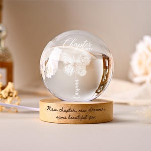 May include: A clear glass sphere with a butterfly and floral design, resting on a wooden base. The sphere has the words "Chapter thirty Caspian" etched on it. The base reads "New chapter, new dreams, same beautiful you."