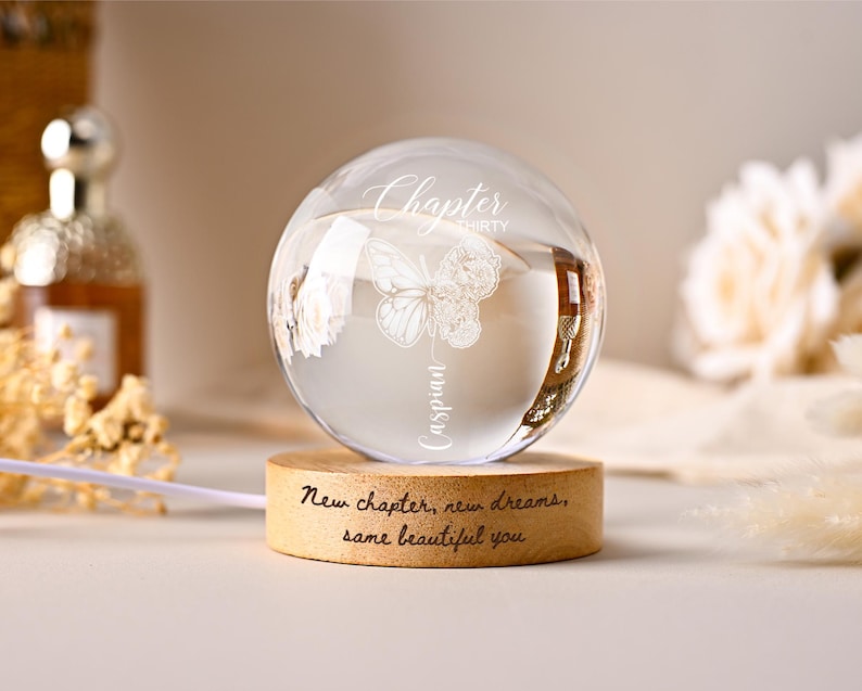 May include: A clear glass sphere with a butterfly and floral design, resting on a wooden base. The sphere has the words "Chapter thirty Caspian" etched on it. The base reads "New chapter, new dreams, same beautiful you."
