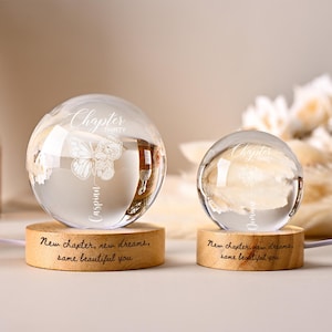 May include: Two clear glass spheres with engraved text and butterfly designs sit on wooden bases. The larger sphere reads "Chapter Thirty Caspian" and the smaller one reads "Chapter Dream". The bases are inscribed with "New chapter, new dreams, same beautiful you."