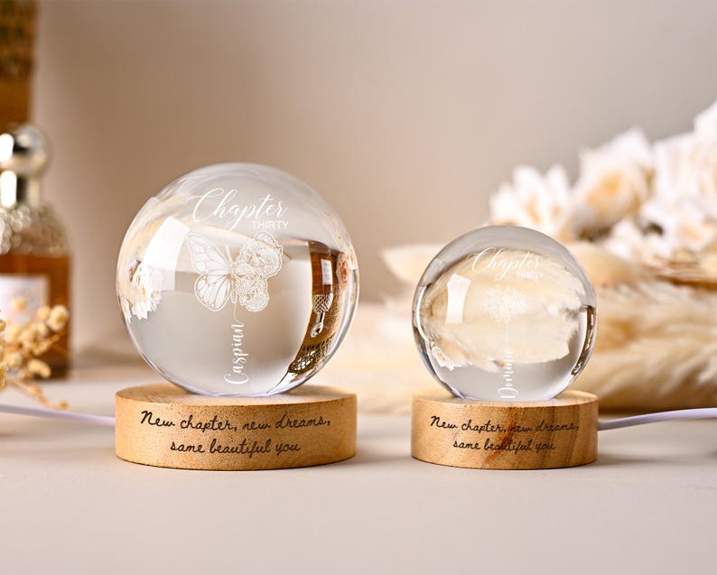 May include: Two clear glass spheres with engraved text and butterfly designs sit on wooden bases. The larger sphere reads "Chapter Thirty Caspian" and the smaller one reads "Chapter Dream". The bases are inscribed with "New chapter, new dreams, same beautiful you."