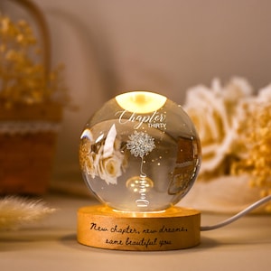 May include: A decorative glass sphere with a warm, glowing light inside, resting on a wooden base. The sphere features a floral design and the words "Chapter Thirty" and "New chapter, new dreams, same beautiful you."