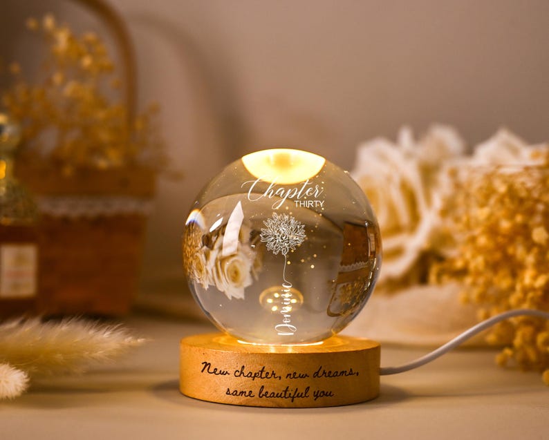 May include: A decorative glass sphere with a warm, glowing light inside, resting on a wooden base. The sphere features a floral design and the words "Chapter Thirty" and "New chapter, new dreams, same beautiful you."