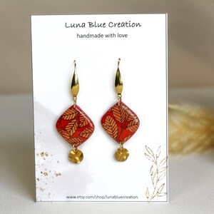 Puede incluir: Un par de pendientes colgantes rojos y dorados con un diseño geométrico. Los pendientes presentan un colgante rojo en forma de diamante con un patrón de hojas doradas y un dije de disco dorado. Los pendientes se muestran en una tarjeta blanca con el texto "Luna Blue Creation handmade with love" y una dirección de sitio web.