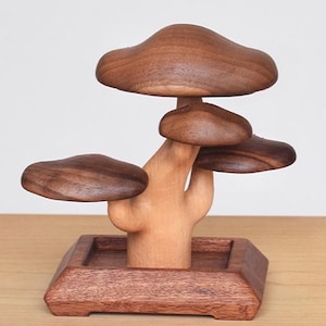 May include: A wooden sculpture of a tree with mushroom-shaped leaves. The tree is made of light and dark brown wood and sits on a square base. The sculpture is on a wooden surface.