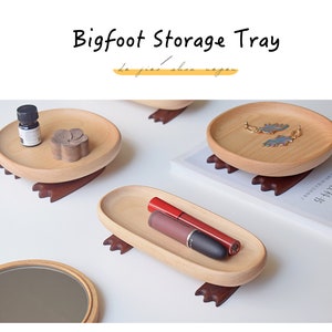 May include: Three wooden trays with small feet, each with different items inside. The first tray has a small bottle and a wooden flower. The second tray has a pair of earrings. The third tray has two tubes of lipstick. The trays are made of light wood and have a natural finish.