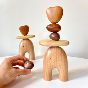 May include: Two wooden sculptures with rounded bases and a heart-shaped top. The sculptures are stacked with wooden circles and a heart-shaped top. The sculptures are made of light brown wood.