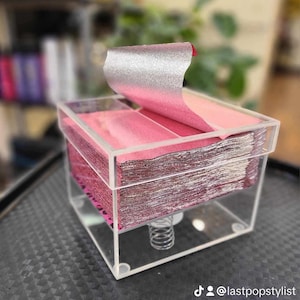 May include: A clear acrylic box filled with stacks of thin, metallic pink and silver foil sheets. The box has a spring mechanism to dispense the foil.