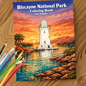 May include: A coloring book titled "Biscayne National Park Coloring Book for Adults" featuring a lighthouse scene with a sunset backdrop. Colored pencils in various colors are arranged on a wooden surface, ready for use.
