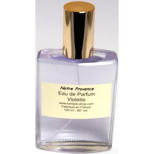 Peut inclure: Un flacon de parfum clair et rectangulaire avec un bouchon doré. Le flacon contient un liquide violet clair et porte une étiquette crème avec le texte "Notre Provence Eau de Parfum Violette". Le flacon est de 100 ml.