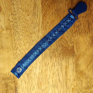May include: A navy blue pacifier clip with a polka dot ribbon design. The clip features a plastic clasp on one end and a metal ring on the other. The ribbon has a white polka dot pattern.