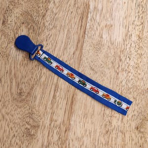 May include: A blue pacifier clip with a car pattern. The clip features a blue plastic clip and a fabric strap with a repeating pattern of colorful cars on a white stripe against a blue background. The clip is approximately 8 inches long.