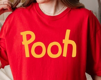May include: A red t-shirt with the word "Pooh" printed in yellow on the front. The shirt is being worn by a person sitting on a bed.