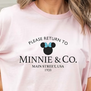 Pu&ograve; includere: Maglietta rosa con una grafica in bianco e nero di Minnie Mouse con un fiocco blu. Il testo recita "Please return to Minnie & Co. Main Street, USA 1928."