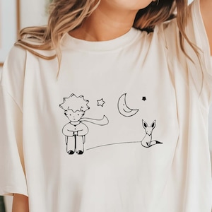 May include: A white t-shirt with a black and white illustration of The Little Prince, a fox, a crescent moon, and stars.