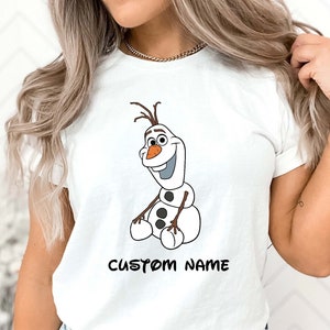 May include: White t-shirt featuring a cartoon illustration of Olaf from Frozen. Olaf is depicted with a carrot nose, twig hair, and black button accents. The text "CUSTOM NAME" is printed in black below the character.