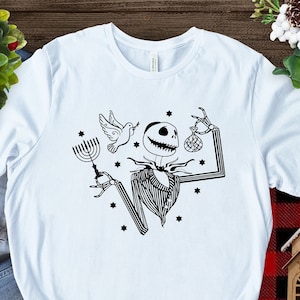 May include: A light blue t-shirt featuring a black and white graphic of a skeleton holding a menorah and a Christmas bauble. The skeleton is wearing a striped shirt and a bow tie. There are stars around the skeleton.