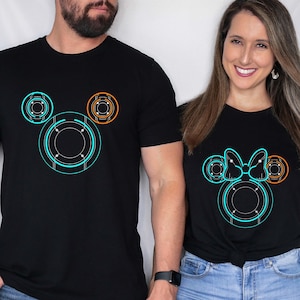 May include: A couple wearing matching black t-shirts with a stylized Mickey Mouse design. The man's shirt features a blue and orange Mickey Mouse head, while the woman's shirt features a blue and orange Mickey Mouse head with a bow.