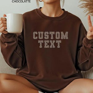 May include: A dark chocolate-colored sweatshirt with the words "CUSTOM TEXT" in a leopard print design. The sweatshirt is a crew neck style. A white mug is held in the left hand.