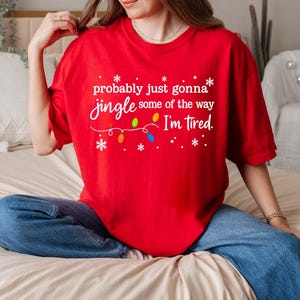 May include: A red t-shirt with the text "probably just gonna jingle some of the way I'm tired" in white script. The design includes snowflakes and a string of colorful Christmas lights. The shirt is paired with blue jeans.
