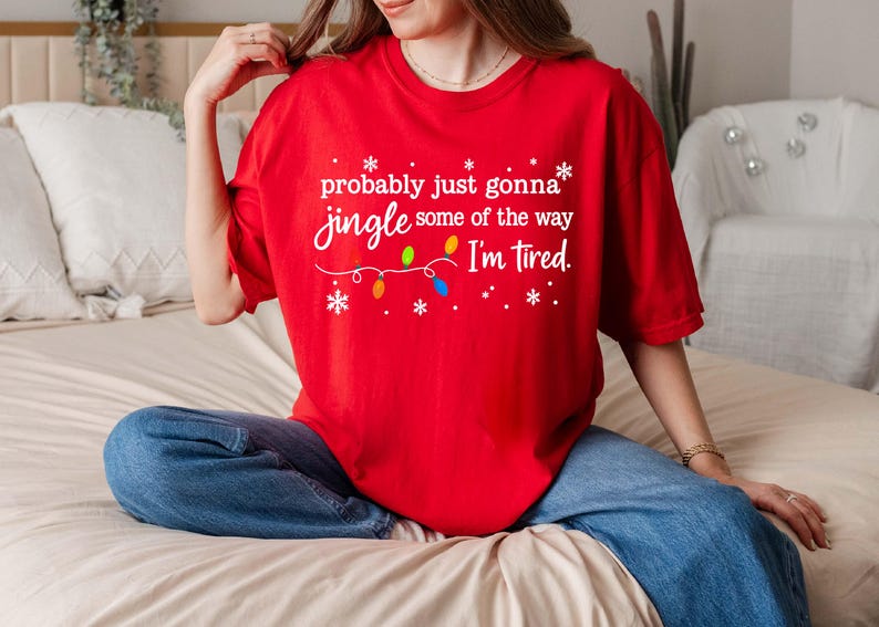 May include: A red t-shirt with the text "probably just gonna jingle some of the way I'm tired" in white script. The design includes snowflakes and a string of colorful Christmas lights. The shirt is paired with blue jeans.