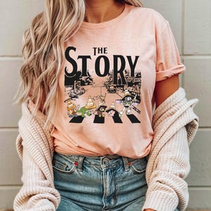 Puede incluir: Camiseta rosa claro con el texto negro "THE STORY OF TOYS". El gr&aacute;fico muestra personajes de dibujos animados cruzando una calle con conos de tr&aacute;fico. La camiseta est&aacute; hecha de un material suave.