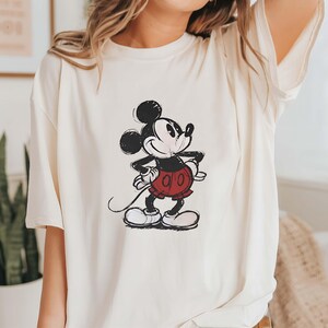 May include: A white t-shirt with a black and white illustration of Mickey Mouse. Mickey is wearing red shorts and is smiling.