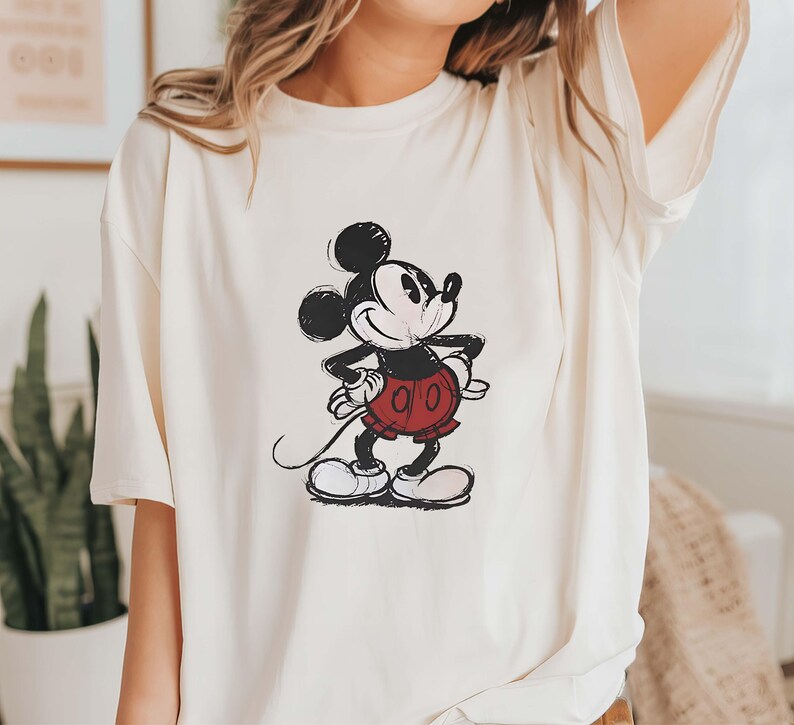 May include: A white t-shirt with a black and white illustration of Mickey Mouse. Mickey is wearing red shorts and is smiling.
