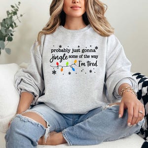 May include: A light gray sweatshirt with the text "probably just gonna jingle some of the way I'm tired." The text is in black with colorful Christmas lights and snowflakes. The sweatshirt is a crew neck style.