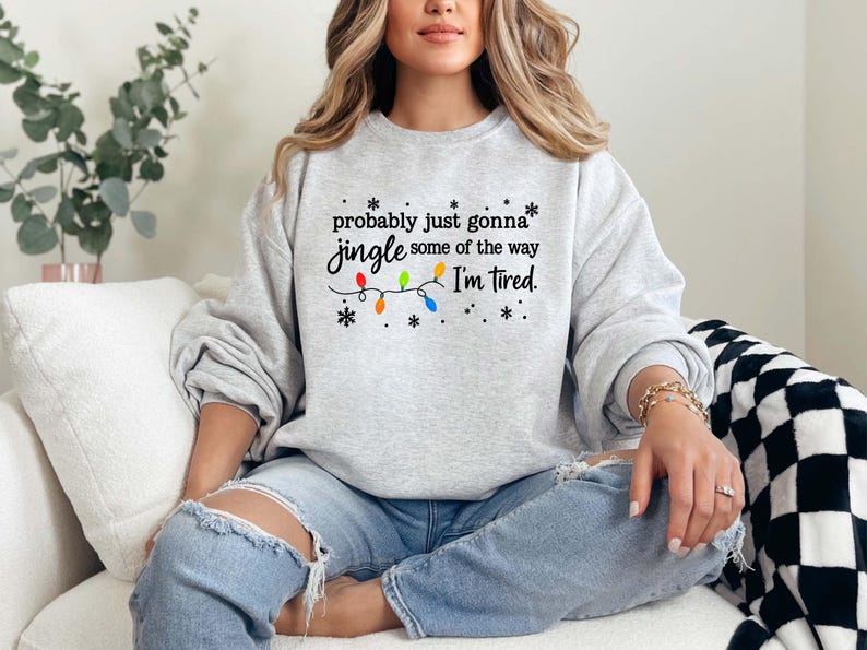 May include: A light gray sweatshirt with the text "probably just gonna jingle some of the way I'm tired." The text is in black with colorful Christmas lights and snowflakes. The sweatshirt is a crew neck style.