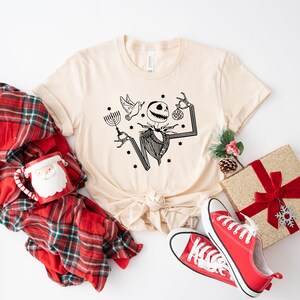 May include: A beige t-shirt featuring a black and white illustration of a skeleton holding a menorah and a bauble. The shirt is surrounded by a red and black checked blanket, a red mug with a Santa face, a red and gold gift box with a red bow, and red trainers.