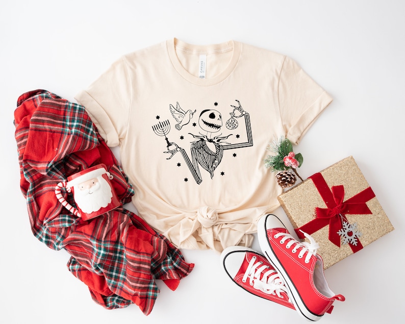 May include: A beige t-shirt featuring a black and white illustration of a skeleton holding a menorah and a bauble. The shirt is surrounded by a red and black checked blanket, a red mug with a Santa face, a red and gold gift box with a red bow, and red trainers.