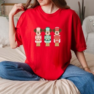May include: A red t-shirt featuring a graphic of three cartoon nutcracker figures. Each figure has a Mickey Mouse-like face and wears a different colored uniform: pink, teal, and red. The shirt is made of a soft material.