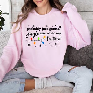 May include: A light pink sweatshirt with the text "probably just gonna jingle some of the way I'm tired." The text is accompanied by a string of colorful Christmas lights and snowflakes. The sweatshirt is a casual, comfortable style.