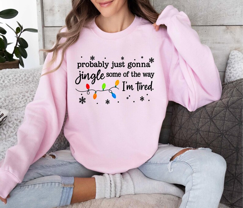 May include: A light pink sweatshirt with the text "probably just gonna jingle some of the way I'm tired." The text is accompanied by a string of colorful Christmas lights and snowflakes. The sweatshirt is a casual, comfortable style.