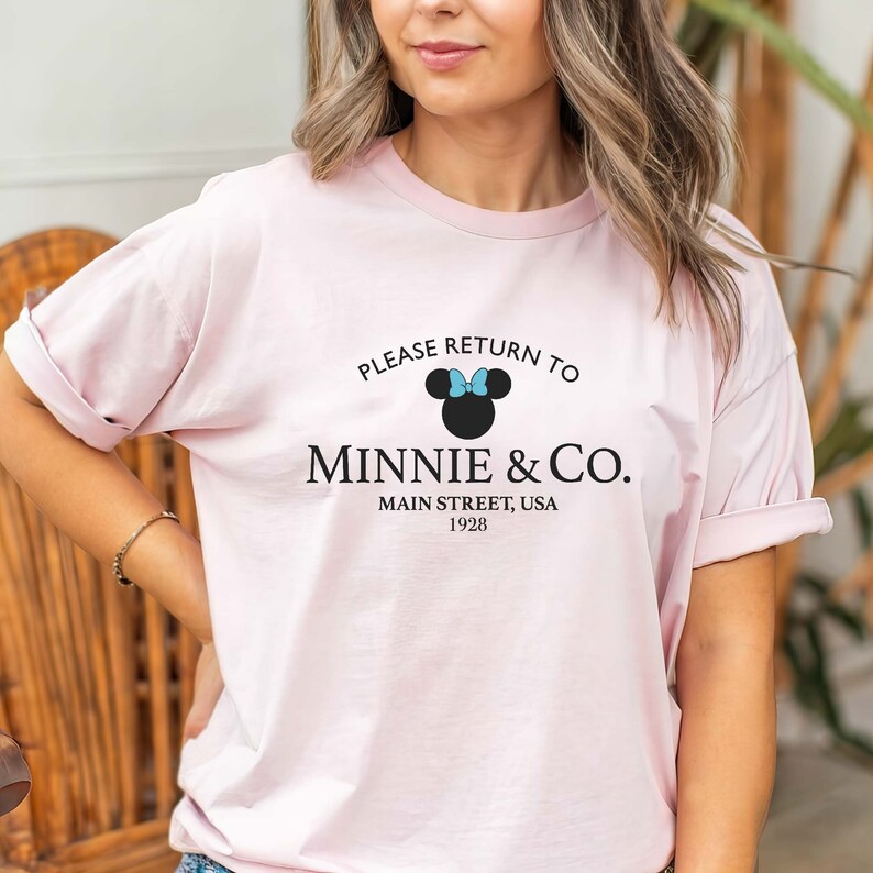 Pu&ograve; includere: Maglietta rosa con una grafica in bianco e nero di Minnie Mouse con un fiocco blu. Il testo recita "Please return to Minnie & Co. Main Street, USA 1928."