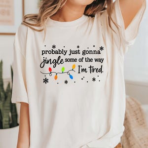May include: Cream-colored t-shirt with the text "probably just gonna jingle some of the way I'm tired." The text is accompanied by a string of colorful Christmas lights and snowflakes.