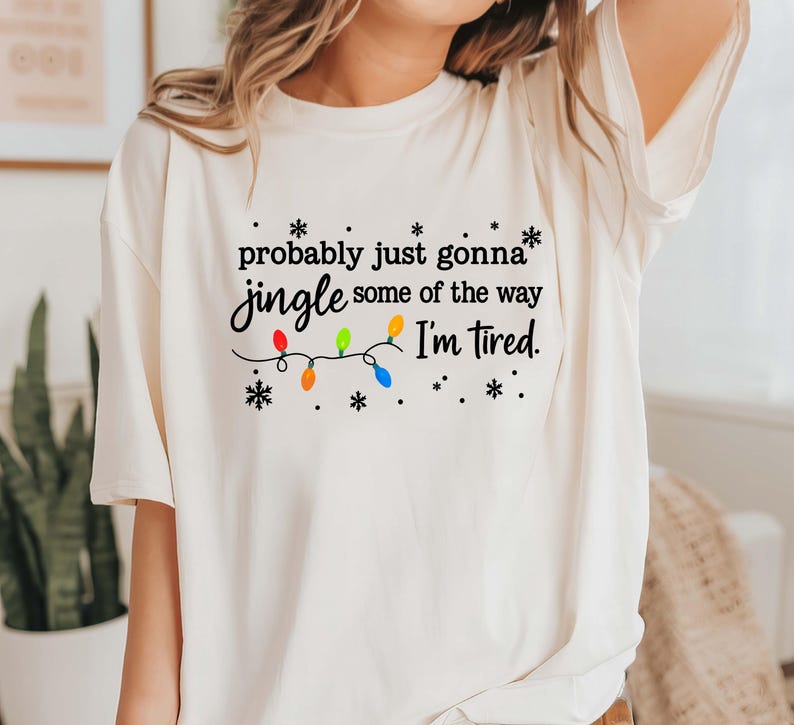 May include: Cream-colored t-shirt with the text "probably just gonna jingle some of the way I'm tired." The text is accompanied by a string of colorful Christmas lights and snowflakes.
