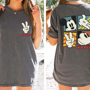 Pode incluir: Uma camiseta cinza com um design vintage do Mickey Mouse. O design apresenta o rosto do Mickey, a m&atilde;o fazendo um sinal de paz e a palavra "Mickey" em vermelho.