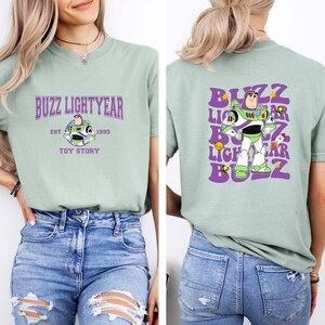 May include: A light green t-shirt with a purple graphic of Buzz Lightyear from Toy Story. The front of the shirt says "Buzz Lightyear Est 1995 Toy Story". The back of the shirt has a repeated graphic of Buzz Lightyear with the text "Buzz Lightyear Buzz" in a retro font.