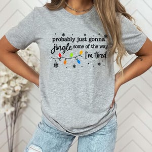 May include: A gray t-shirt with the text "probably just gonna jingle some of the way I'm tired." The text is in black script with colorful Christmas lights and snowflakes.