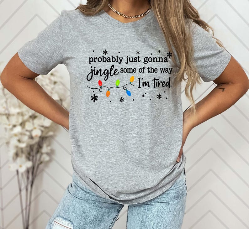 May include: A gray t-shirt with the text "probably just gonna jingle some of the way I'm tired." The text is in black script with colorful Christmas lights and snowflakes.